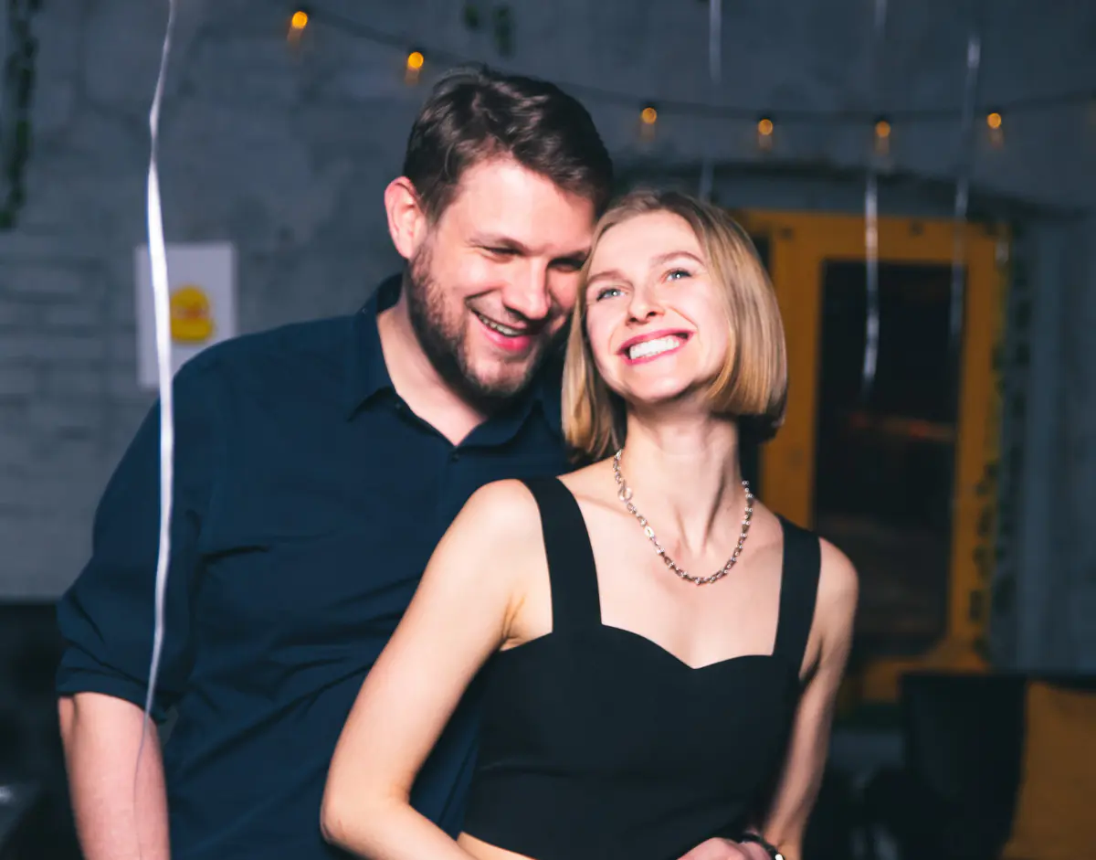Мужчина нежно обнимает улыбающуюся блондинку в черном платье на фоне теплых ретро гирлянд