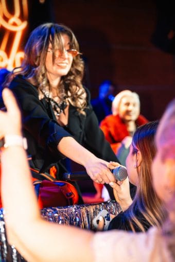 Фотография женщины в полумраке, готовой к выступлению, с микрофоном и черным платьем.